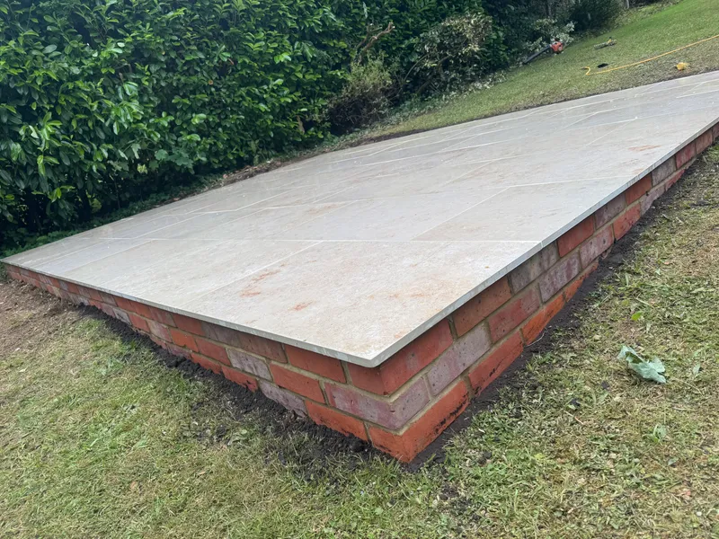 Raised base faced in red brick and topped with pale porcelain paving, beside a hedge and lawn