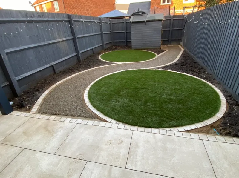 Circular lawns edged in light block paving, linked by a curving gravel path edged to match, beside a porcelain patio