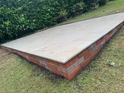 Raised base faced in red brick and topped with pale porcelain paving, beside a hedge and lawn