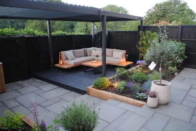 The same Hazlemere garden after a design and build makeover, with grey paving, a raised planter and a dark deck with outdoor sofas under a louvred pergola
