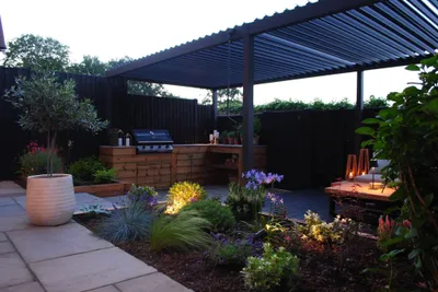 The finished Hazlemere garden at dusk, with lit borders and an outdoor kitchen with a built-in barbecue beneath the pergola