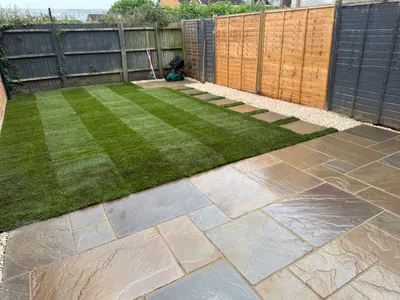 New striped turf lawn beside a sandstone patio, with a stepping-stone path and fencing behind