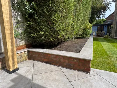 Pale paved patio framed by a brick raised bed with stone coping, leading to a striped lawn