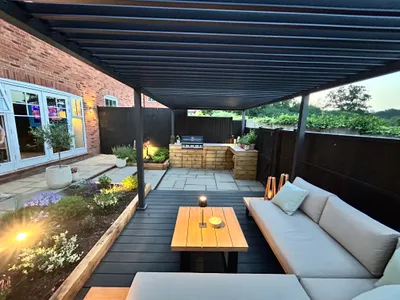 Dark louvred aluminium pergola covering a deck with outdoor sofas, next to the house and an outdoor kitchen