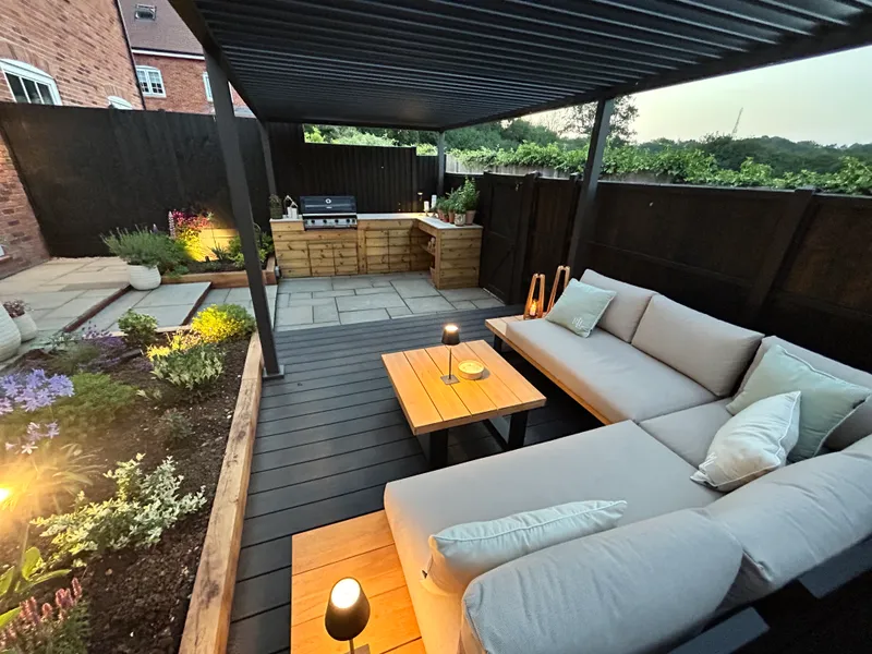 Outdoor seating area on a dark deck beneath a louvred pergola, with an outdoor kitchen with a built-in barbecue beyond