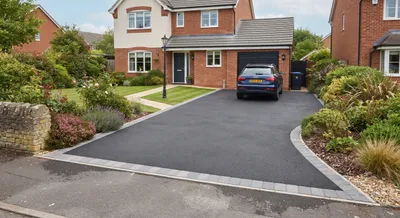 Black tarmac driveway with a grey block paved edge, leading to a brick house and garage, with planted borders on both sides